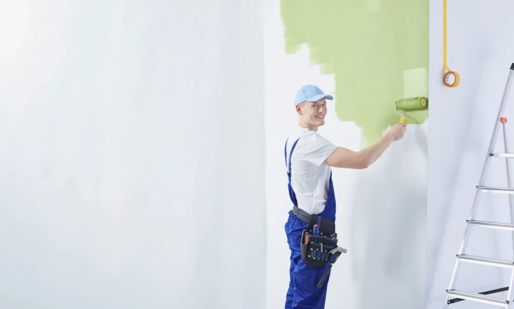 Handwerker trägt Farbe mit Rolle auf Wand auf, daneben Leiter in leerer Wohnung